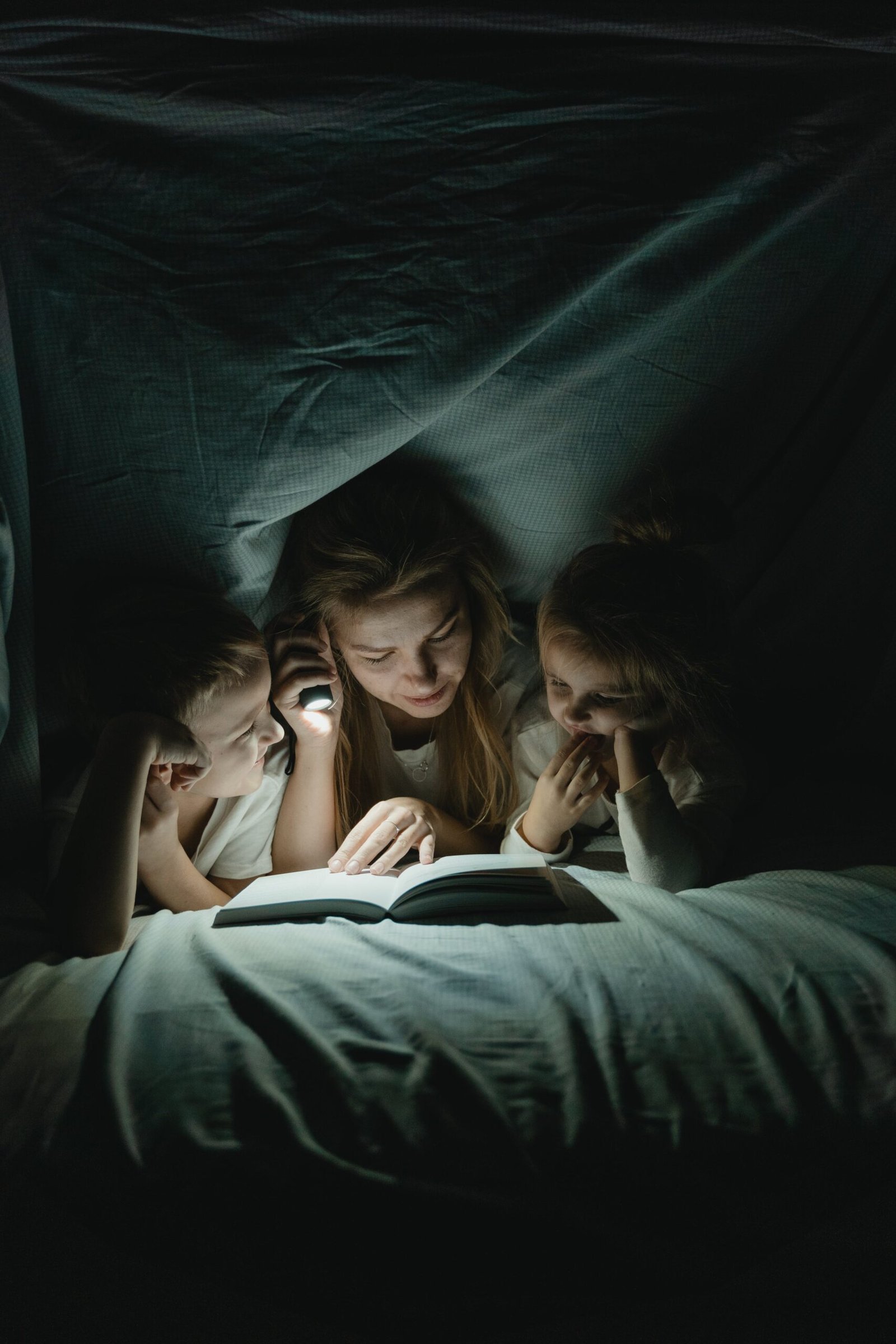 A woman is telling her two kids a bedtime story using a flashlight to read.